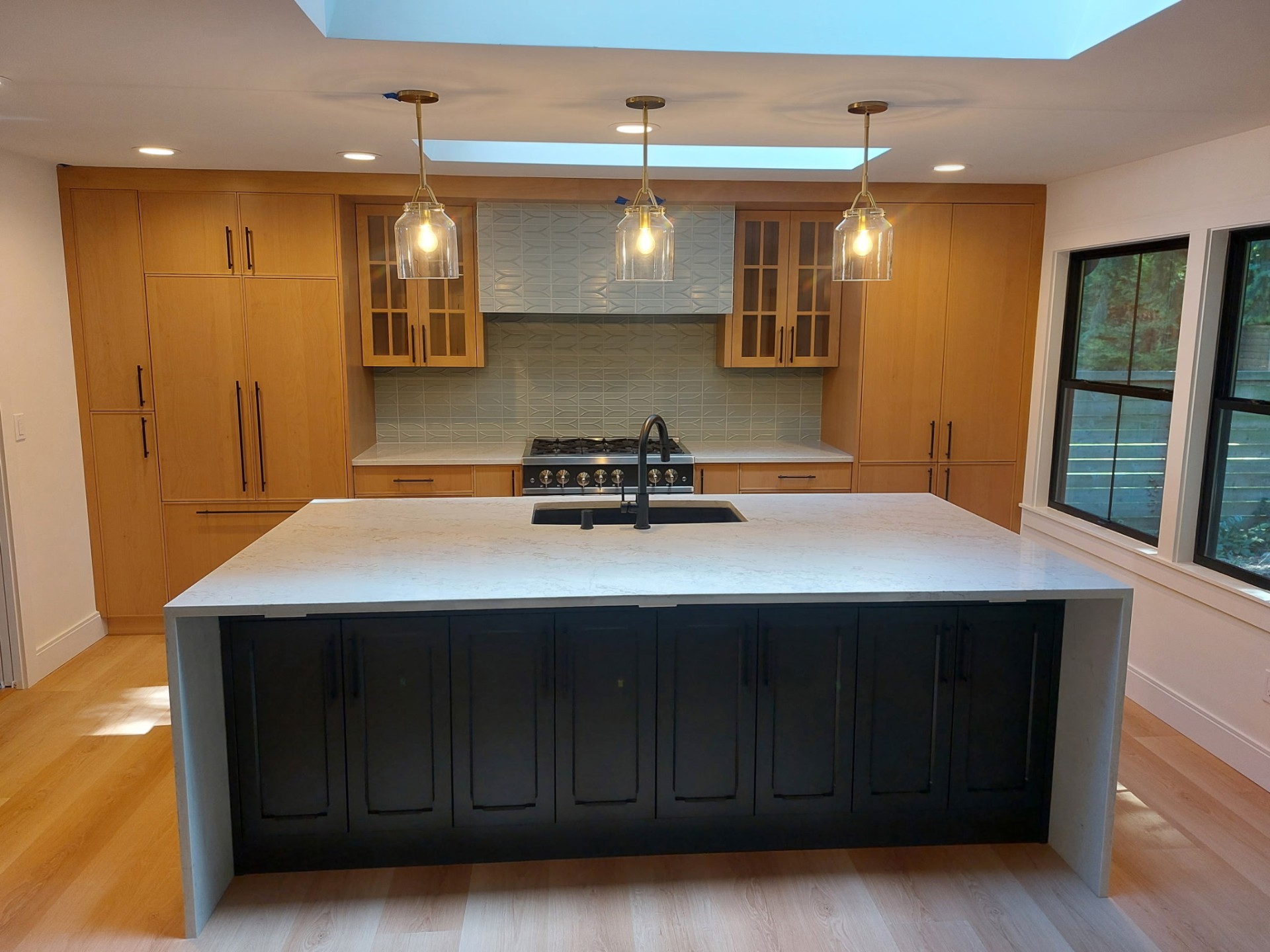 Quartz waterfall island with black cabinetry, warm wood cabinets, brass pendants and skylights — Seattle area kitchen